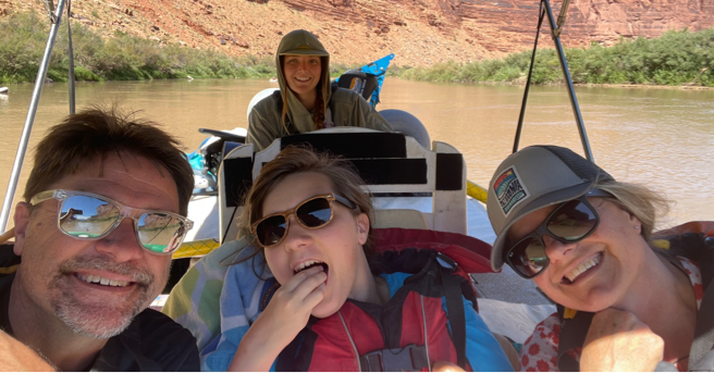 Lucy on a boat, smiling and putting her hand in her mouth, in between her parents. A boat driver is smiling in background.