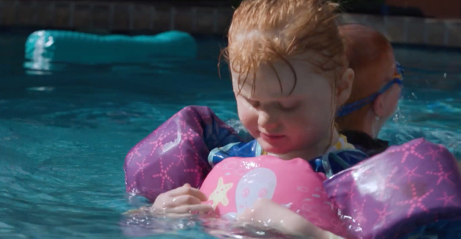 Minna in floaties looking down into the pool water. Her brother, Oscar, is behind her.
